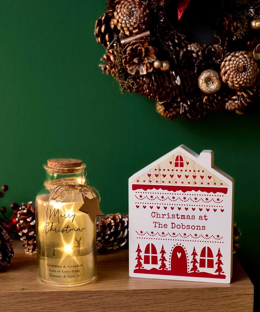 Personalised Christmas lantern and house-shaped decoration displayed on a wooden table with festive pinecones and a Christmas wreath. Personalised Christmas lantern and house-shaped decoration displayed on a wooden table with festive pinecones and a Christmas wreath.